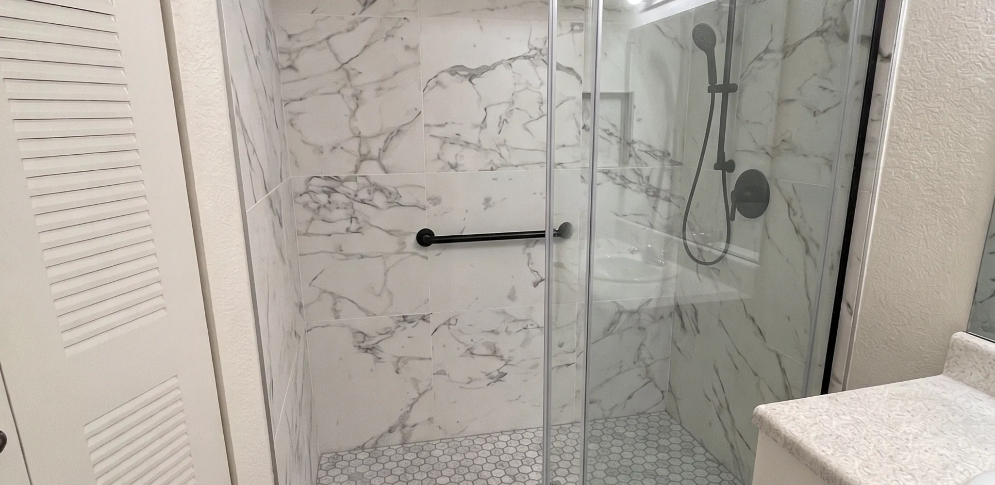 Walk-in shower with a low curb, a matte black grab bar, a hand shower on a slide bar and a two-panel sliding glass door
